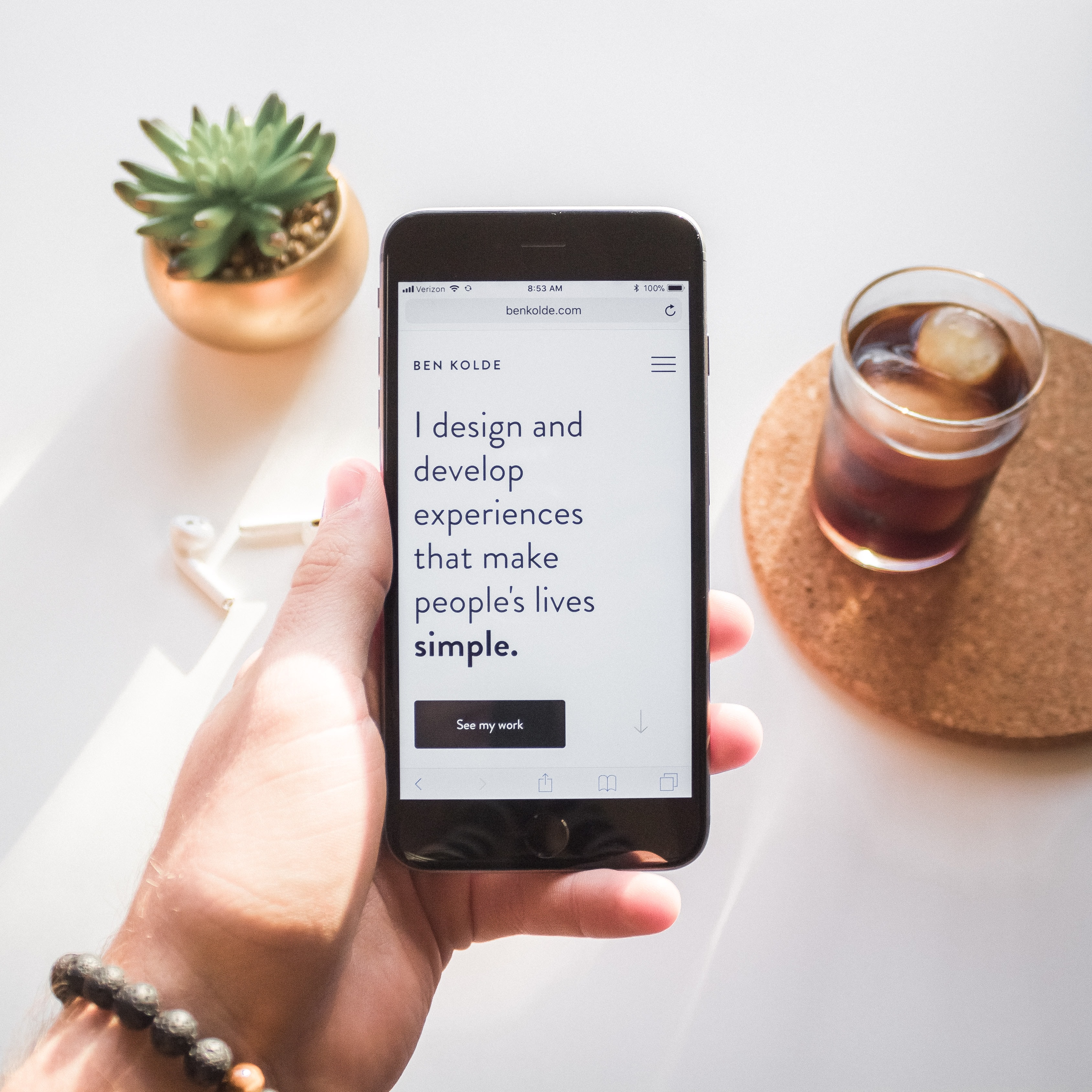 Hand holding an iPhone with a quote from Ben Kolde 'I design and develop experiences that make people's lives simple'. There is a plant, glass of coffee, and airpods in the background.